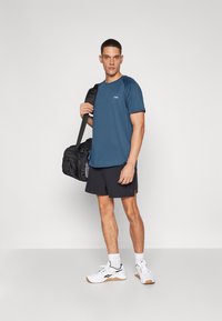 Outfit sportivo con una maglietta blu a maniche corte, pantaloni corti neri e sneakers bianche. Il modello tiene una borsa da palestra nera. Design pulito e vestibilità casual.