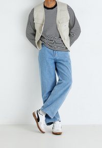 Gilet senza maniche beige sopra una camicia a maniche lunghe a righe bianco e nere, abbinato a jeans blu chiaro e scarpe da ginnastica bianche con dettagli neri.