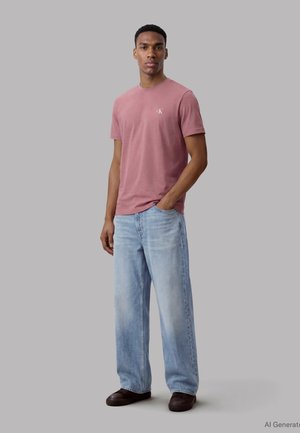 Homme debout, main dans la poche, portant un t-shirt à manches courtes rose poussiéreux, un jean large bleu clair et des chaussures marron foncé, sur un fond gris neutre.
