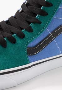 Baskets en daim vert et toile bleue avec lacets noirs, semelle blanche et bande latérale noire. Détails de couture visibles le long des bords.