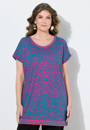 Kvinde iført en løs, blågrøn og pink zebrastribet kortærmet top med store gyldne hoop-øreringe, står foran en hvid væg.