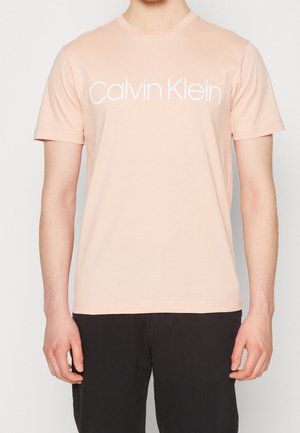 Personne portant un t-shirt à manches courtes rose clair avec le texte blanc "Calvin Klein" sur la poitrine et un pantalon noir, debout devant un fond uni.