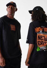 T-shirt nere oversize, una con il logo arancione "NEW ERA" sul petto, l'altra con una grafica che rappresenta un muro di mattoni e elementi urbani sul retro.