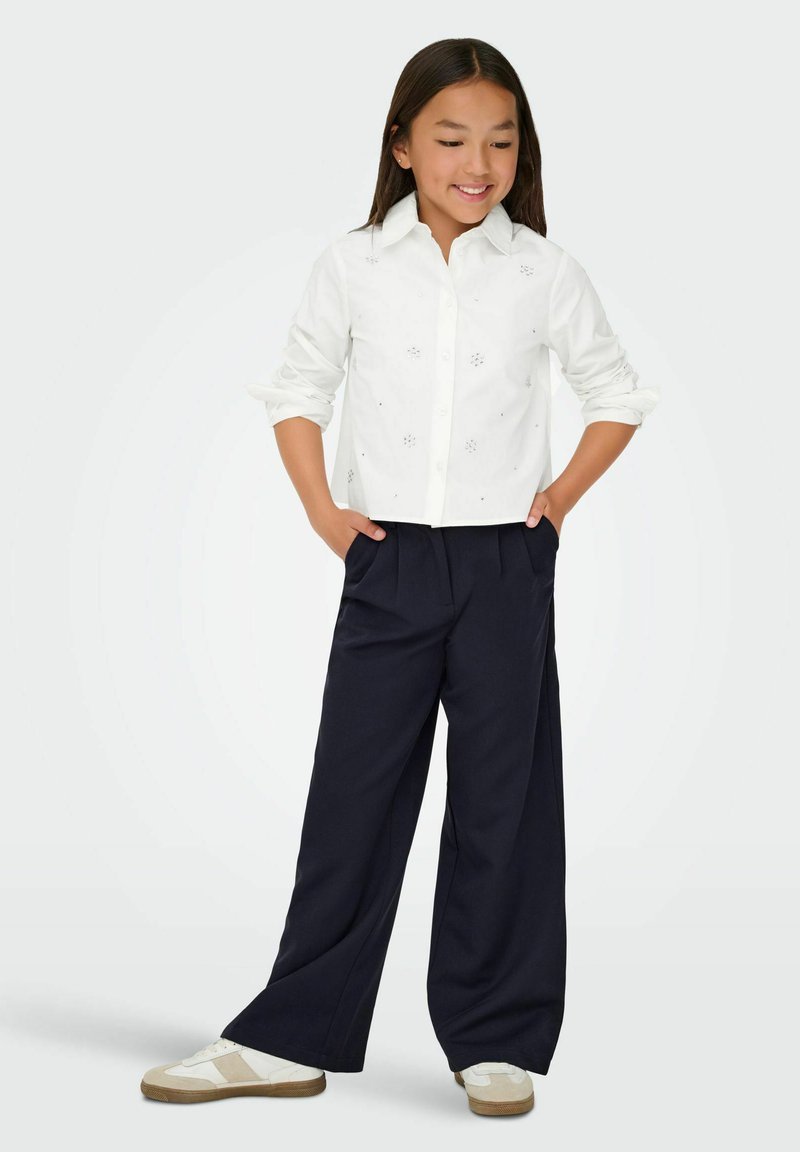 Camicetta bianca con dettagli ricamati, abbinata a pantaloni larghi blu navy e sneakers bianche con accenti color tan.