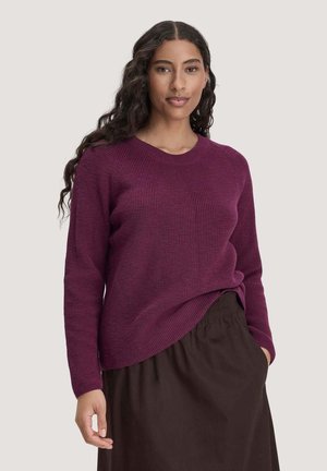 Vrouw met lang krullend haar, gekleed in een paarse gebreide trui en een donkerbruine rok, staand tegen een eenvoudige lichte achtergrond.