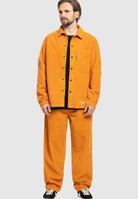 Orange corduroy jacket with black buttons, matching corduroy pants, and a black shirt underneath; features a front pocket with multicolored accents.
