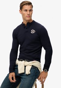 Marineblaues Langarm-Poloshirt mit knöpfbarem Kragen und gesticktem Logo. Getragen mit einem hellen Pullover, der in der Taille gebunden ist, und Jeans.