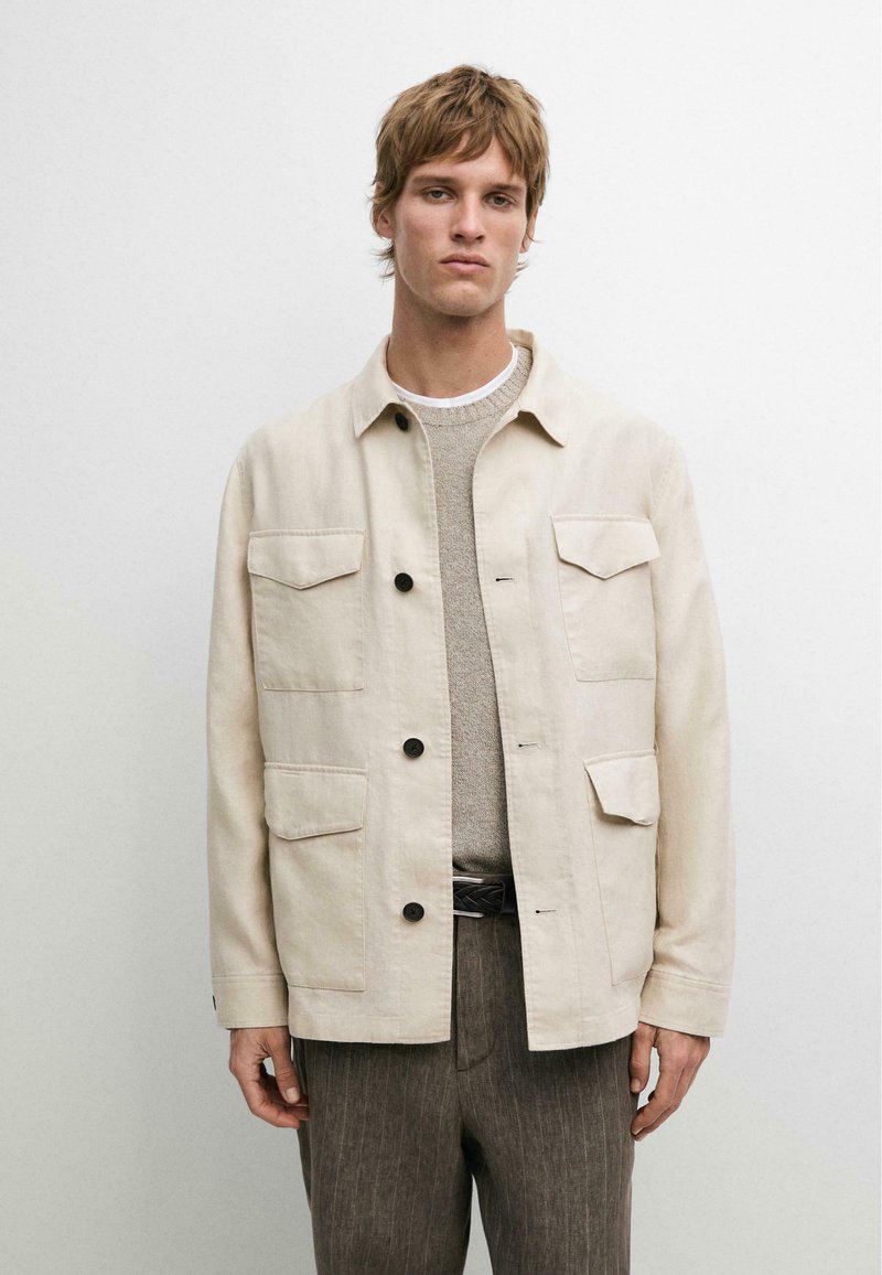 Junger Mann trägt eine beige Jacke mit Knöpfen über einem hellgrauen Pullover und braune Nadelstreifenhose, stehend vor einem schlichten weißen Hintergrund.