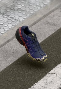 Navy blue trail running shoe with orange accents, a textured sole, and a secure lacing system. Features a cushioned collar and distinct tread design.