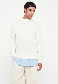 Bărbat îmbrăcat cu un pulover alb din tricot peste o cămașă cu dungi albastre și albe, asortat cu pantaloni largi de culoare crem, stând în fața unui fundal alb.