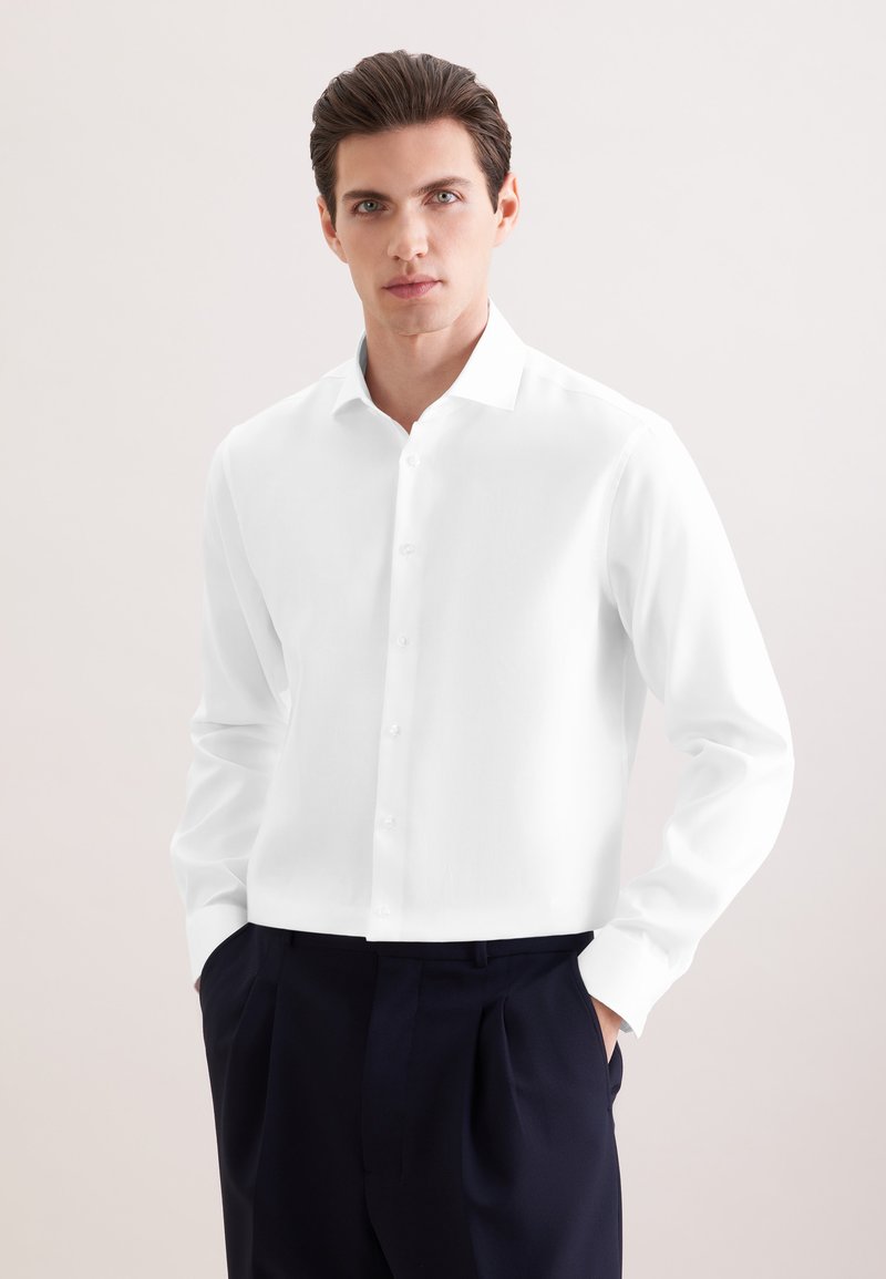 Homme aux cheveux foncés portant une chemise blanche boutonnée et un pantalon sombre, debout les mains dans les poches devant un fond clair uni.