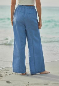 Pantaloni largi din in albastru, cu talie elastică, având două buzunare în spate și o textură netedă, purtați pe o plajă nisipoasă.
