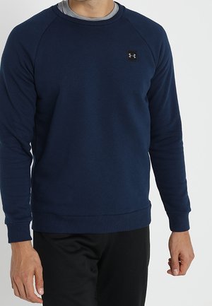 Homme portant un sweat-shirt à manches longues Under Armour bleu marine par-dessus un sous-vêtement gris, associé à un pantalon noir.