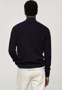 Marineblauer, gerippter Strickpullover mit hohem Kragen, langen Ärmeln und taillierter Passform. Über weißen Hosen getragen, die eine strukturierte Oberfläche zeigen.