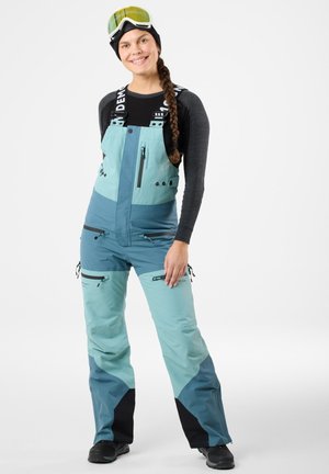 Ski bib w jasnym i ciemnym odcieniu teal, z czarnymi akcentami, regulowanymi ramiączkami, wieloma zapinanymi na zamek kieszeniami oraz luźnym krojem zapewniającym swobodę ruchów.