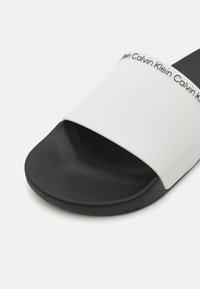 Sort slide sandal med en tekstureret sål og et bredt hvidt bånd med mærkenavnet i sort tekst langs den øverste kant.