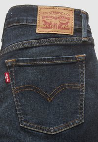 Buzunar posterior alblugilor Levi's de culoare albastru închis, cu cusături portocalii, etichetă roșie Levi's și patch de brand din piele bej pe talie.