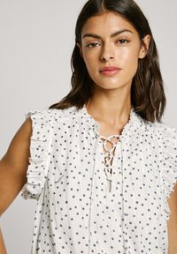 Blusa bianca con volant e pois blu navy, scollatura con lacci e texture delicata. Presenta un sottile logo vicino all'orlo.