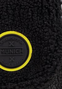 Tela negra texturizada con un parche de goma negra circular con borde amarillo, que muestra "MUNICH" y detalles de marca adicionales.