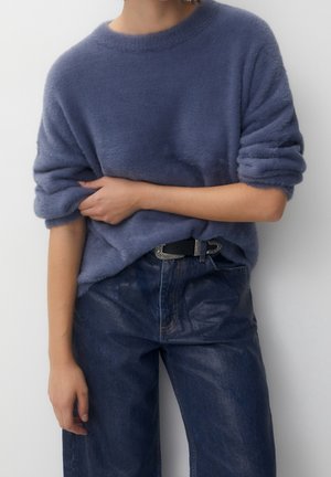 Personne portant un pull bleu doux avec les manches retroussées et un jean en denim bleu foncé avec une ceinture noire, debout devant un fond uni.