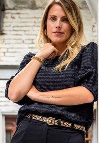 Zwarte textuurblouse met pofmouwen, gecombineerd met hoge taille broek en een riem met gouden accenten. Gouden armband om de pols.