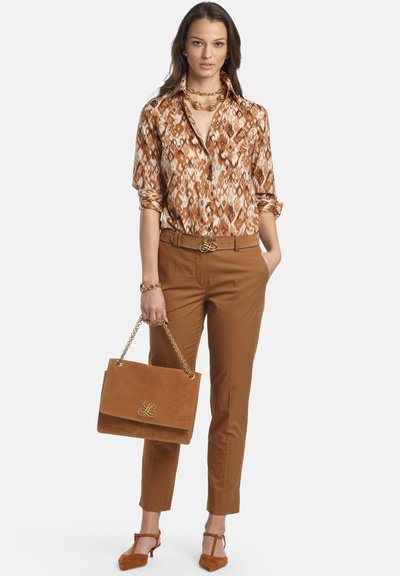 Blusa a maniche lunghe con fantasia in toni caldi, abbinata a pantaloni sartoriali color cammello. Accessori includono una collana a catena d'oro, una cintura e una borsa.