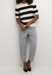 Femme portant un haut à rayures noires et blanches à manches courtes, un pantalon léger gris sur mesure et des escarpins noirs à bout pointu sur fond uni.