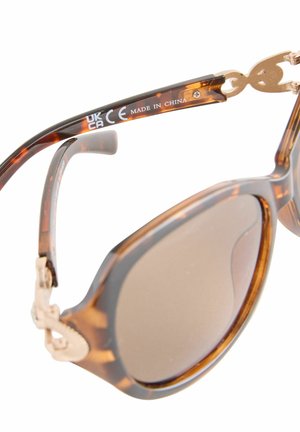 Balinės akiniai nuo saulės su rudais lęšiais, turintys auksinės spalvos detalių ir unikalaus vyrių dizaino. Rėmas turi blizgų paviršių ir apvalią formą.