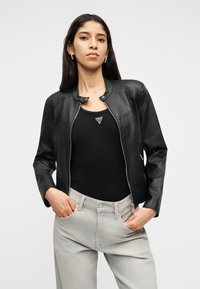 Γυναίκα με μακριά σκούρα μαλλιά, που φορά ασημένια σκουλαρίκια κρίκους, μαύρο δερμάτινο μπουφάν με φερμουάρ, μαύρη μπλούζα χωρίς μανίκια και ανοιχτό γκρι τζιν, με τα χέρια στις τσέπες.