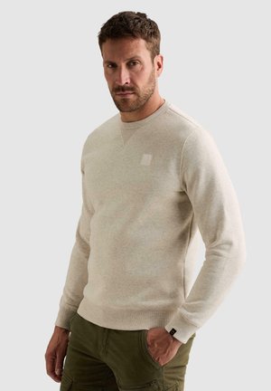 Homme aux cheveux courts et à la barbe, portant un pull beige clair à col rond et un pantalon vert olive, une main dans la poche, sur un fond uni.