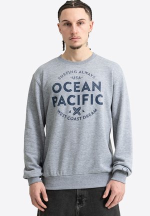 Mężczyzna z warkoczami, ubrany w jasnoszary sweter z logo surfingu "Ocean Pacific" i czarne spodnie, stojący na białym tle.