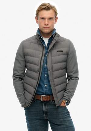 Grijze pufferjas met een gestructureerd ontwerp, voorzien van een ritssluiting aan de voorkant en een knoopsluiting, over een blauwdenim overhemd, gecombineerd met een donkere spijkerbroek.