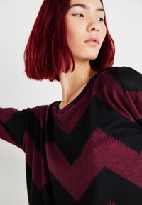 Schwarzer und burgunderfarbener Pullover mit einem Zickzackmuster, aus weichem Material und in lässiger Passform, mit rundem Halsausschnitt und langen Ärmeln.