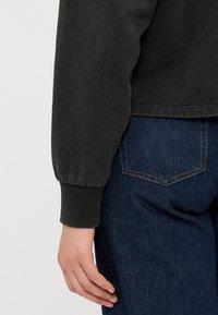 Mørkegrønn genser med puffermer og ribbede mansjetter, parret med mørkeblå denimjeans med synlige sømmer på baklommene.