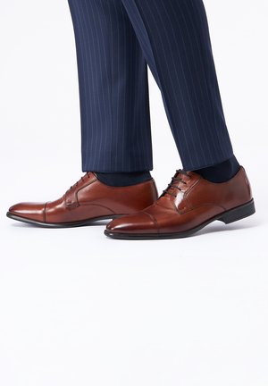 Chaussures habillées en cuir marron avec un bout pointu, une finition texturée et des détails de couture, associées à un pantalon bleu marine à rayures.