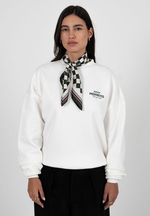 Vrouw met lang donker haar draagt een witte sweatshirt met de tekst "PROHIBITED Racing Club", een zwart-wit geblokte sjaal en een zwarte broek.