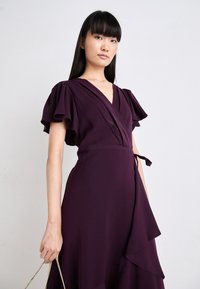 Rochie purpurie dintr-un material moale, cu mâneci fluturate, decolteu în V și un accent de talie legată. Fusta cu straturi adaugă o textură fluidă.