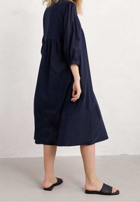 Vestido azul marino, hasta la rodilla, con mangas abullonadas y cintura fruncida, hecho de tejido ligero. Lleva sandalias negras tipo slide.