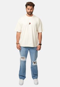Cremefarbenes Oversized-T-Shirt mit gesticktem Design auf der Brust, kombiniert mit hellblauen kaputten Jeans und weißen Sneakers.