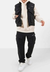 Schwarze gesteppte Weste mit Reißverschlusstaschen über einem cremefarbenen Hoodie, kombiniert mit schwarzen Cargohosen mit Klapptaschen und weißen Sneakern.