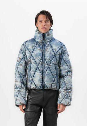 Man wearing a blue patterned quilted puffer jacket with a high collar and black leather pants, standing against a white background.