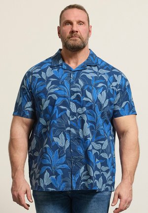 Muskelbepackter Mann mit Bart, der ein blaues kurzärmeliges Hemd mit großem Blattmuster trägt, steht vor einem schlichten hellen Hintergrund.