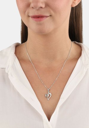 Zilveren hart hangerketting met een kleinere binnenste hart, versierd met heldere accenten, aan een fijne ketting, gedragen over een wit shirt.