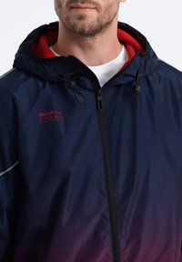 Marineblauwe windbreakerjack met een gradient die overgaat in rood. Voorzien van een frontzak met rits, een verstelbare capuchon en een rode binnenvoering.