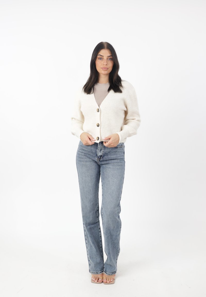 Cardigan crema con scollo a V, texture a coste e cinque bottoni marroni, abbinato a jeans azzurri a vita alta e sandali trasparenti.