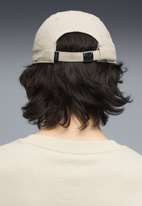 Casquette de baseball beige en coton, dotée d'une sangle arrière avec boucle métallique et d'un logo brodé. Portée avec un t-shirt de couleur claire.