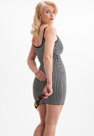 Blonde woman in black-and-white checkered dress holding sunglasses behind her back, looking over her shoulder.