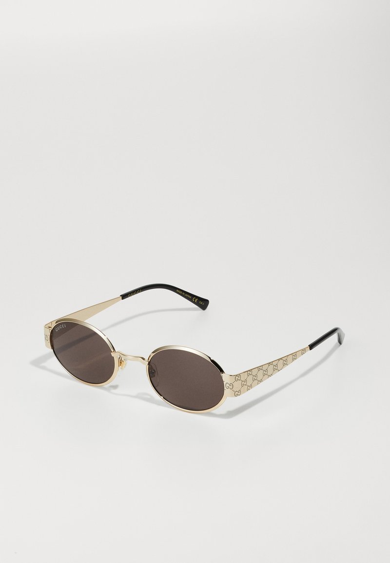 Gafas de sol ovaladas marrones con marco de metal en tono dorado. Patrón decorativo dorado en los laterales, con puntas de patilla de plástico negro. Las lentes ofrecen protección UV.