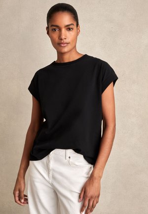 Woman with dark hair tied back wears black short-sleeve shirt and white pants, standing against a plain beige background.