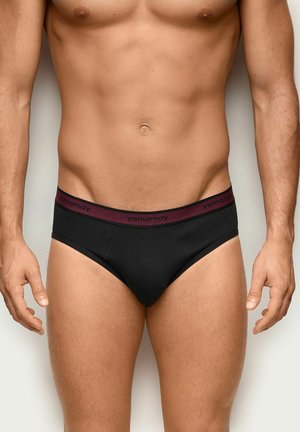 Männlicher Oberkörper trägt schwarze Slips mit einem bordeauxfarbenen Bund mit der Marke "yamamay," und steht vor einem schlichten hellen Hintergrund.
